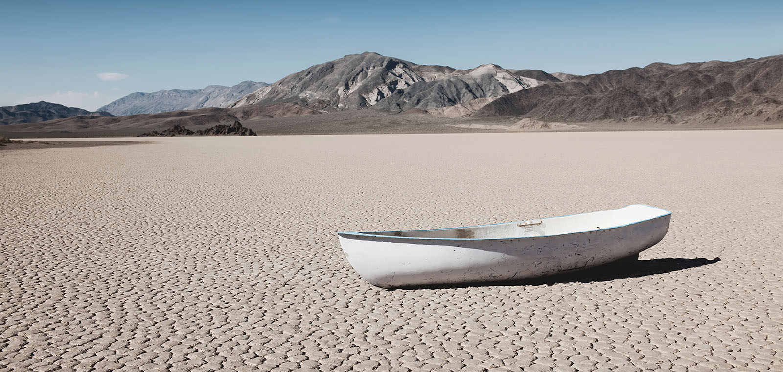 A lone white wooden rowboat rests stranded on a dry, cracked lakebed, contrasting the brown and black parched landscape.