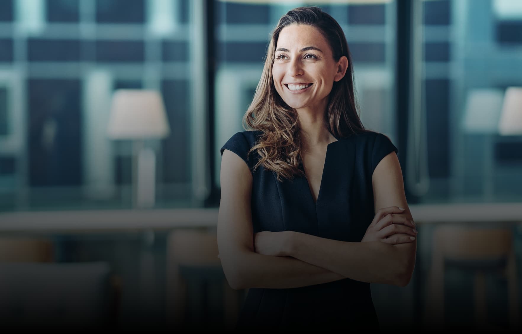 Professional younger woman wearing a navy top in an office with windows in the background, looking to the left with her arms folded.