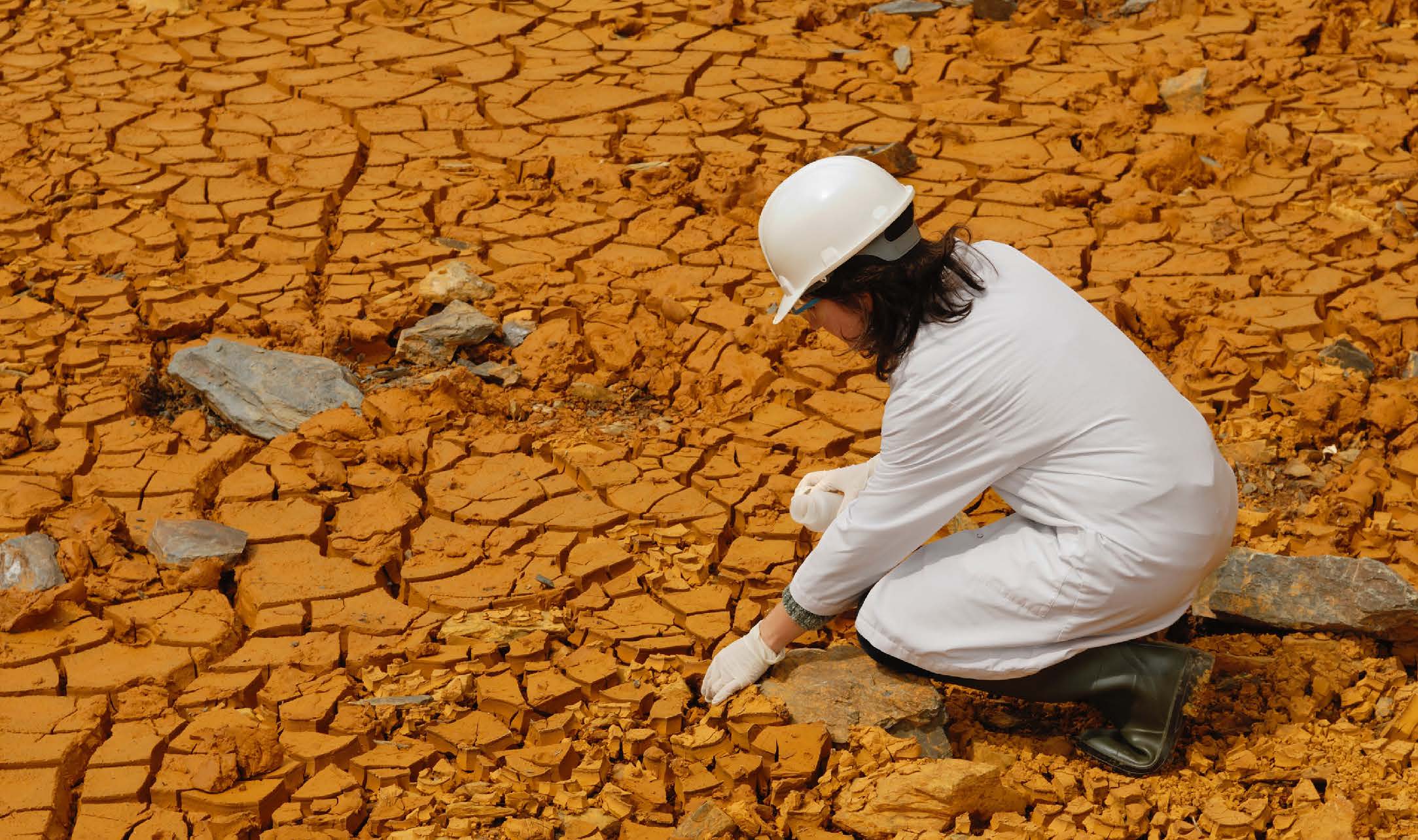 A scientist, dressed in white with a hard hat, kneeling down to inspect the broken earth 