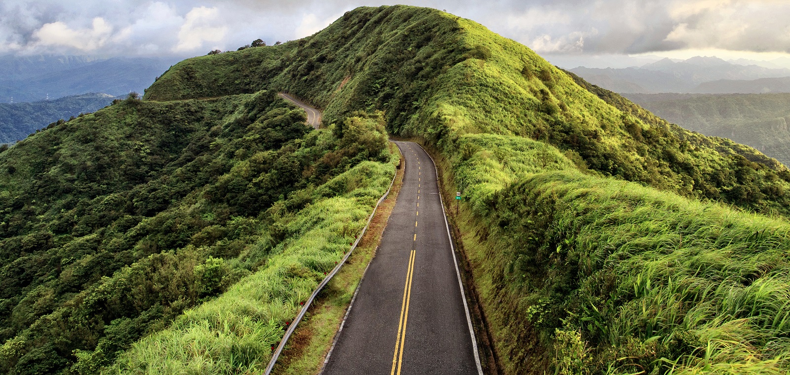 An uneven mountain roadway sharply winds through lush greenery against a cloudy sky