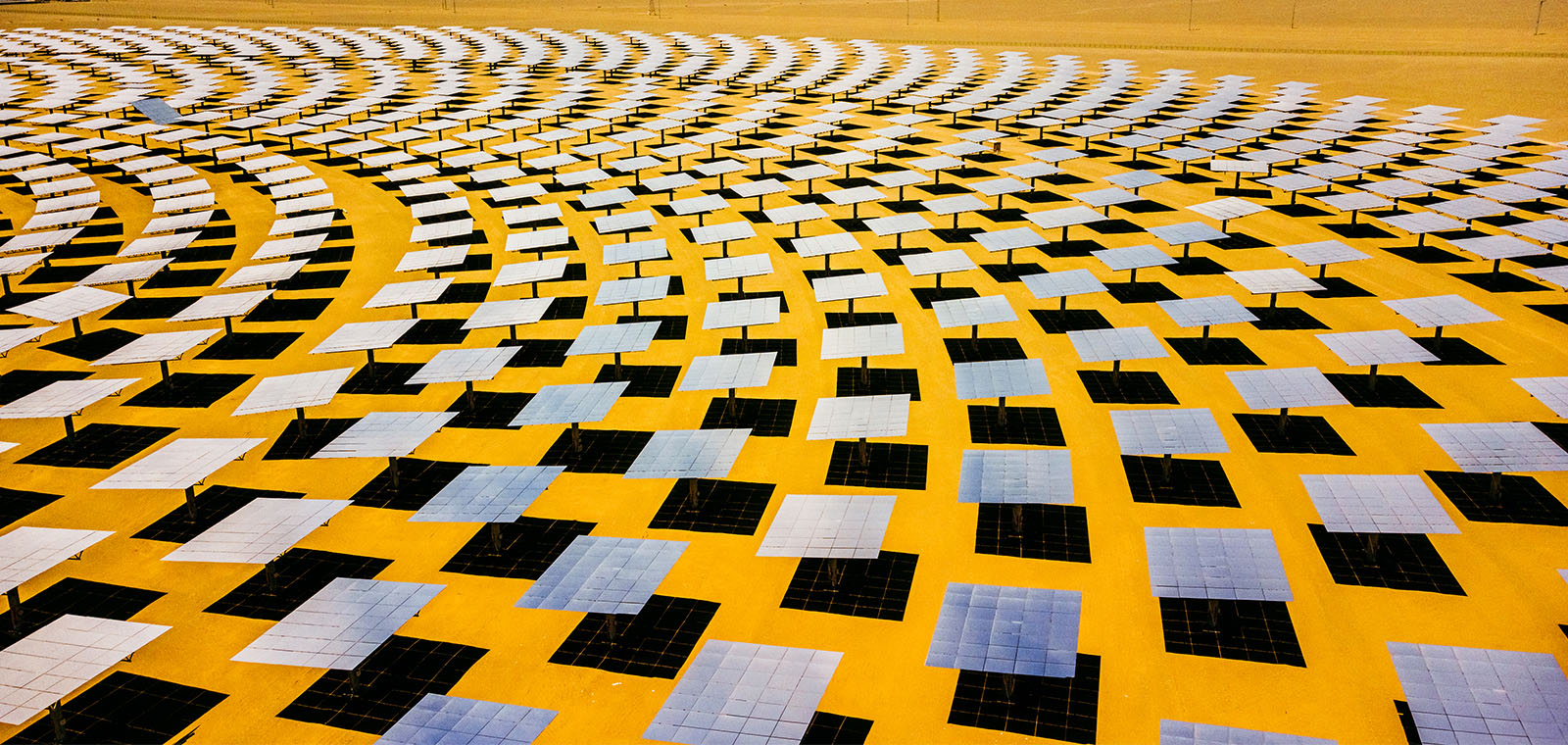A sea of solar power panels stretch out in a curved shape against a yellow desert landscape.