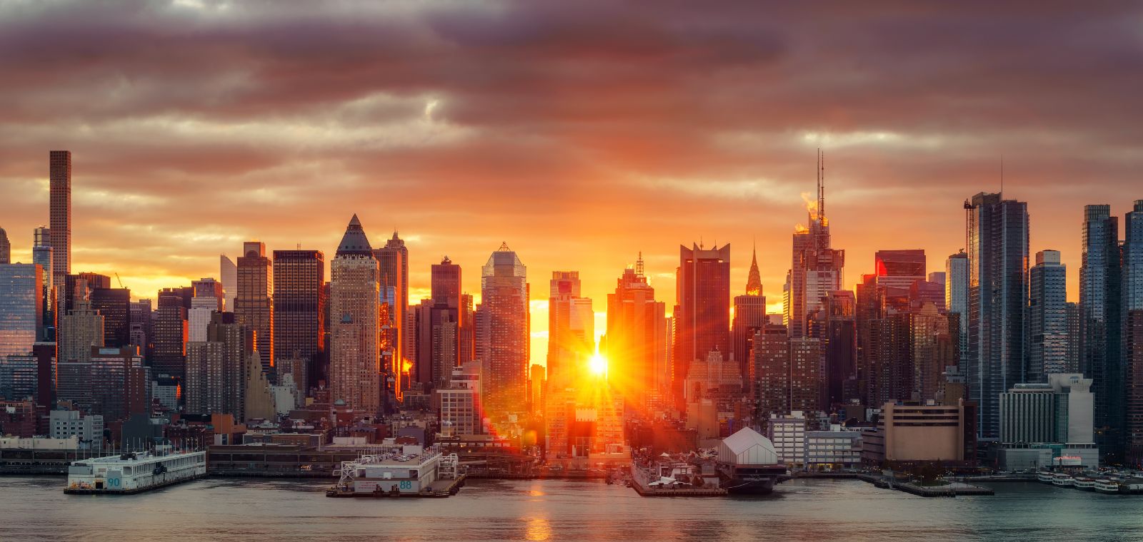 Manhattan at dawn, looking over the East River. The rising sun peeks between skyscrapers under a dark, cloudy sky.