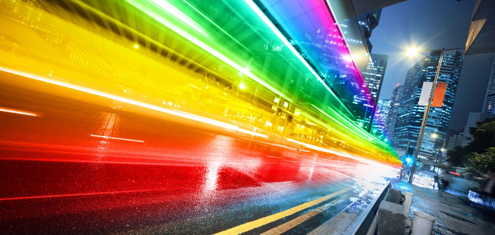 A rainbow-like spectrum of light is seen on a city street, as if the lights of moving cars have been captured in motion. 