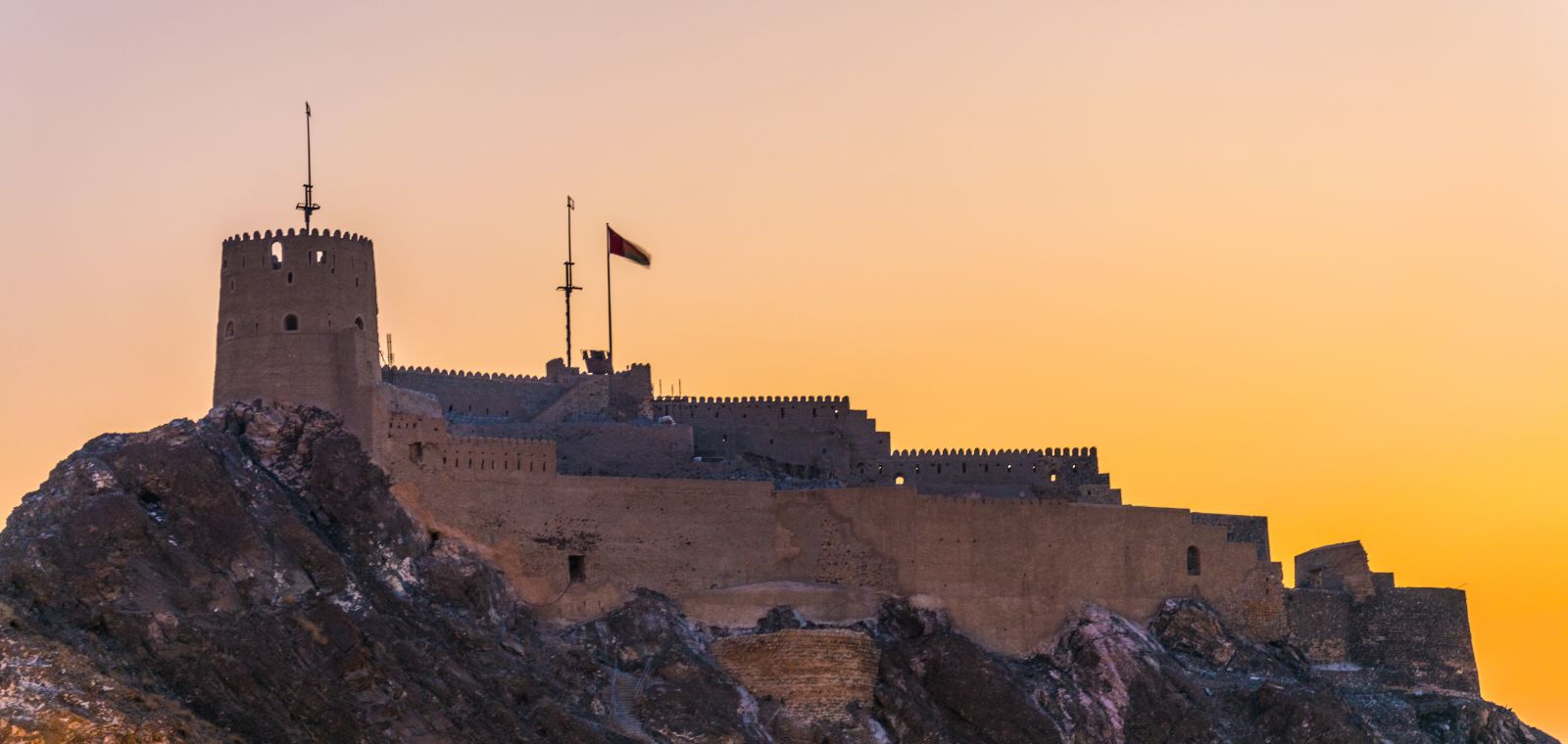 An ancient fortress set high on solid bedrock, with a banner waving above it, against a brilliant yellow sunrise..