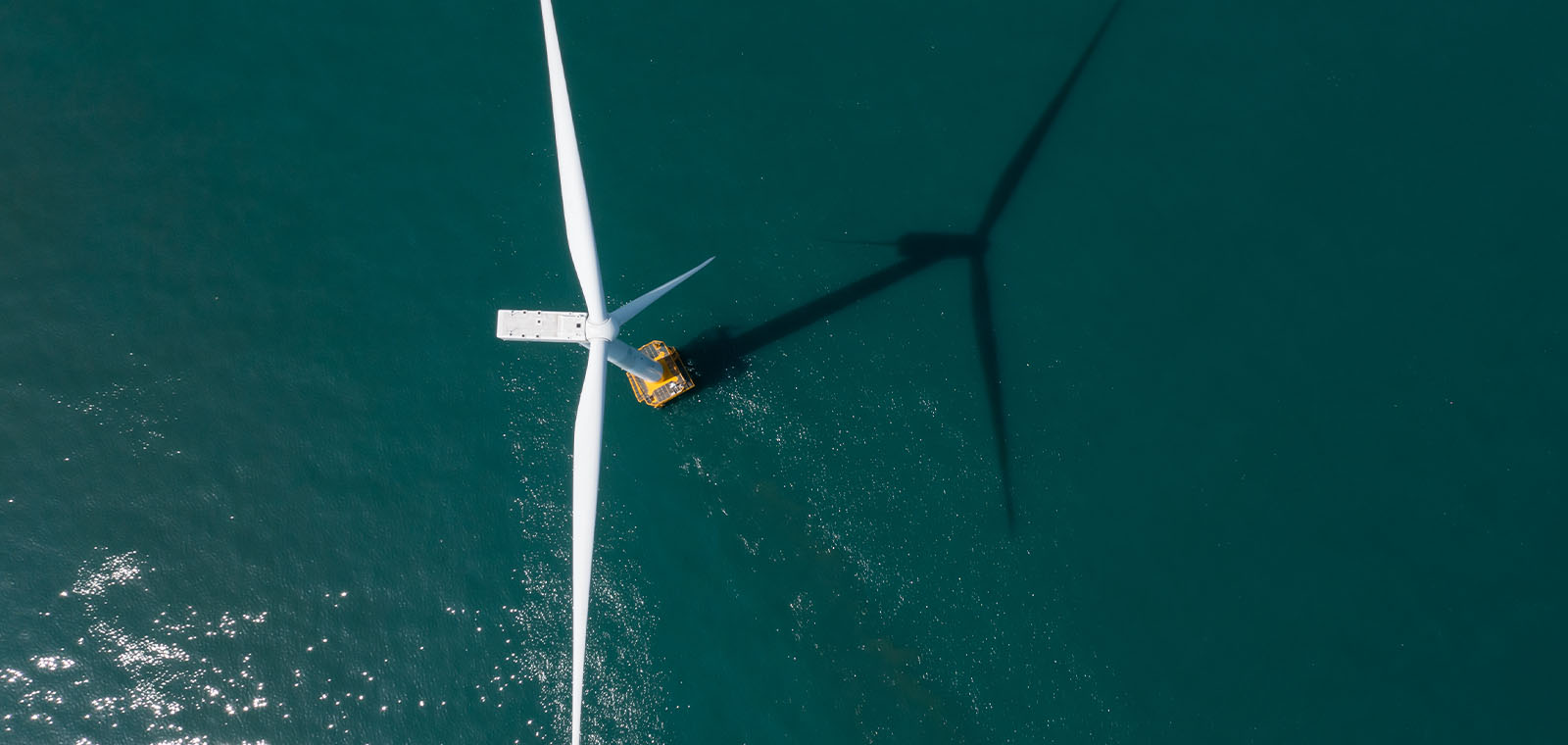 A wind turbine, viewed from above, casts a shadow on the ocean.