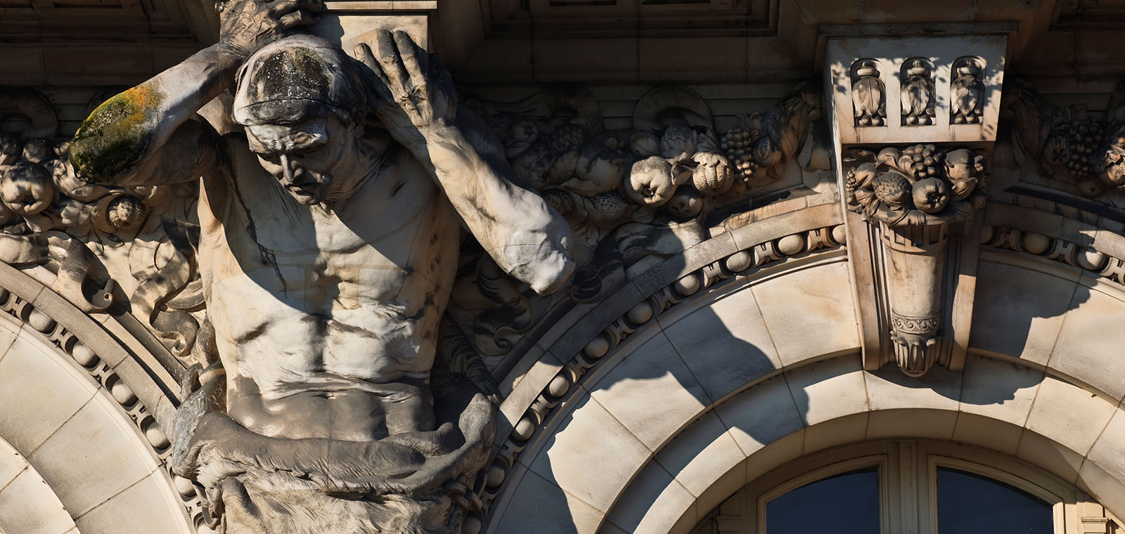 The muscular torso, head, and arms of a statuary Atlas support the entablature of a stone building.