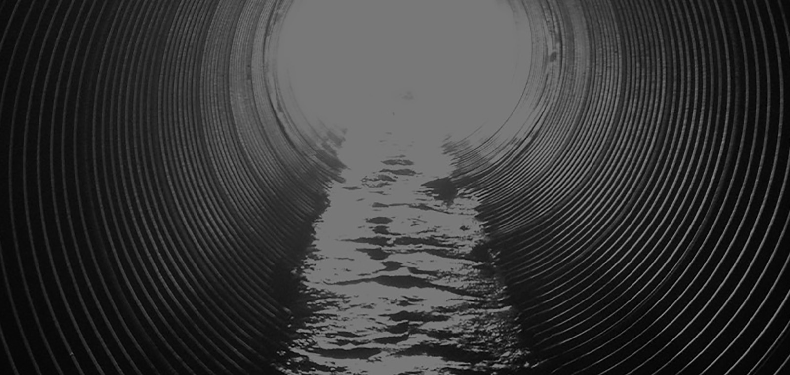 Looking through a corrugated drainage tunnel partially filled with flowing water toward a bright light at its opening.