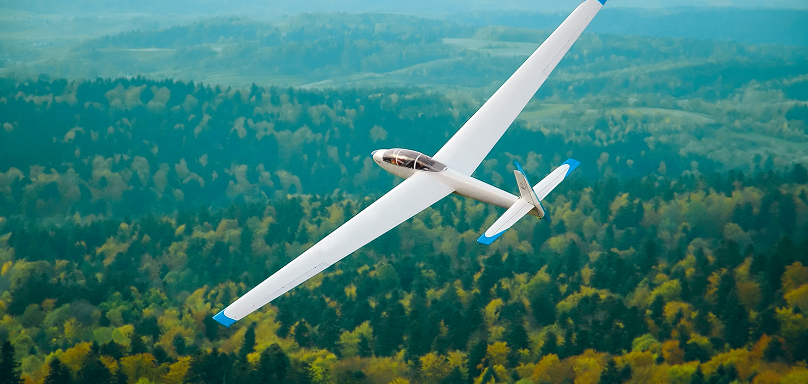 The long, graceful wings of a white glider tilt as the aircraft banks against a background of rolling hills and forests.
