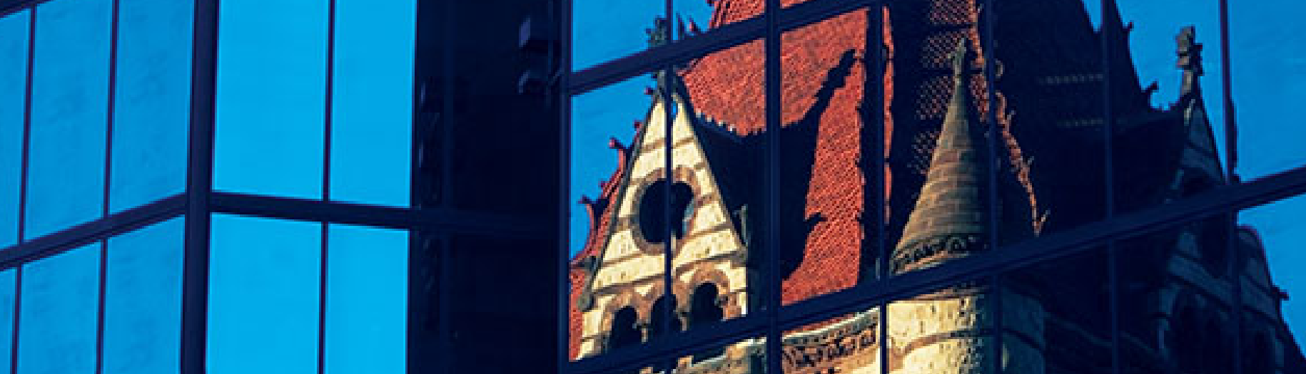 An old, Gothic-style building reflected in the windows of a new office building